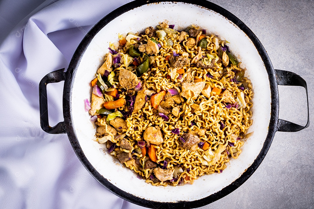 Yakisoba japonês com macarrão frito em panela metálica branca na mesa com toalha de mesa branca e fundo preto, com pauzinhos de comida japonesa... Close view.