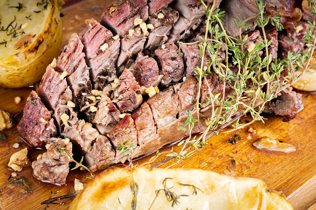 Carne para churrasco. Carne assada com batatas e tomilho numa tábua de madeira, totalmente cortada às rodelas. Vista do topo.