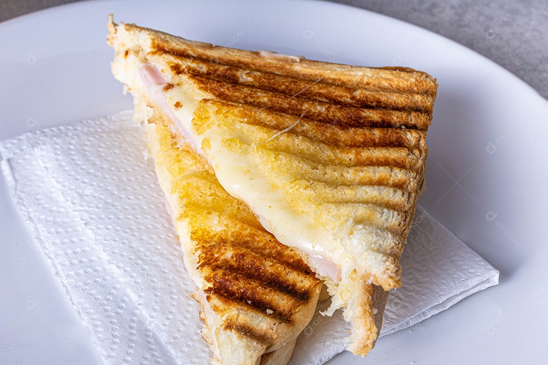 Misto quente. Sanduíche com queijo e presunto na grelha. Servido em toalha xadrez. Acompanha suco de laranja. Visão de perspectiva.