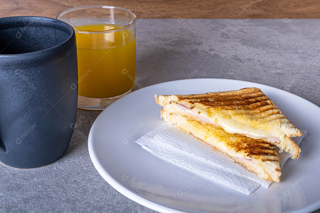 Misto quente. Sanduíche com queijo e presunto na grelha. Servido em toalha xadrez. Acompanha suco de laranja. Visão de perspectiva.