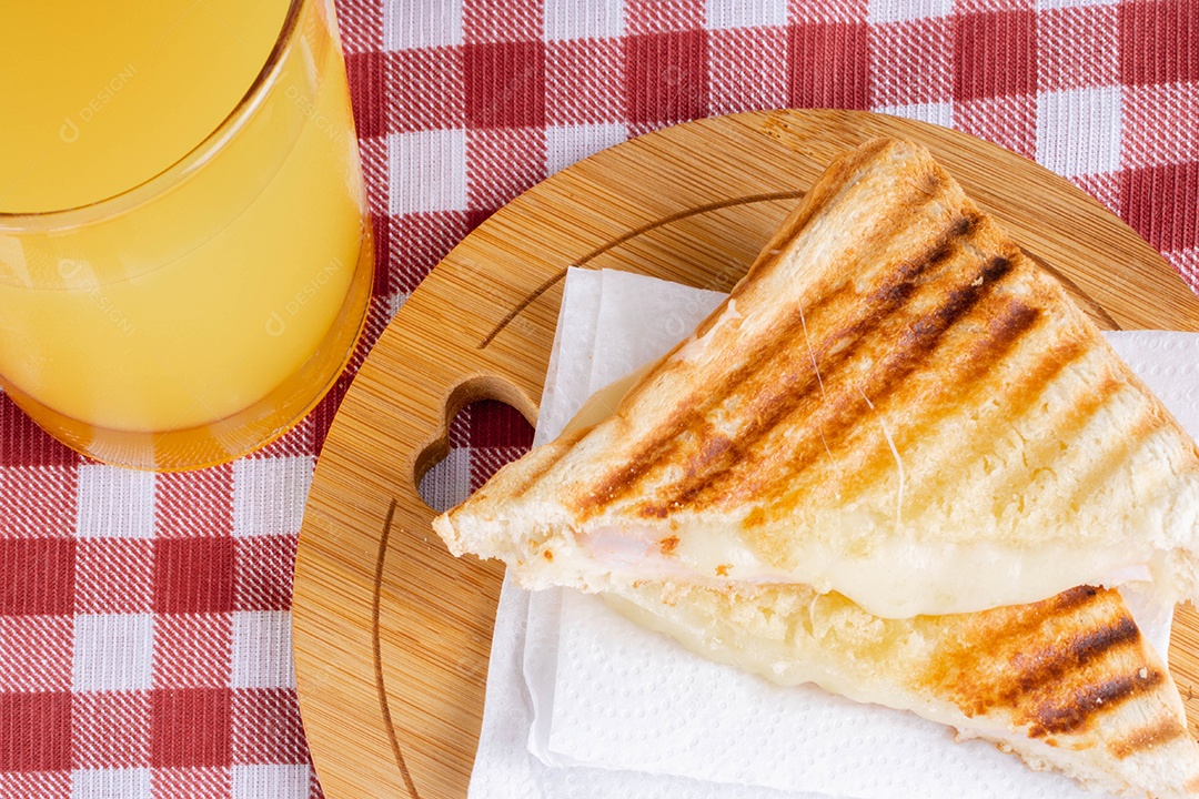 Misto quente. Sanduíche com queijo e presunto na grelha. Servido em toalha xadrez. Acompanha suco de laranja. Visão de perspectiva.