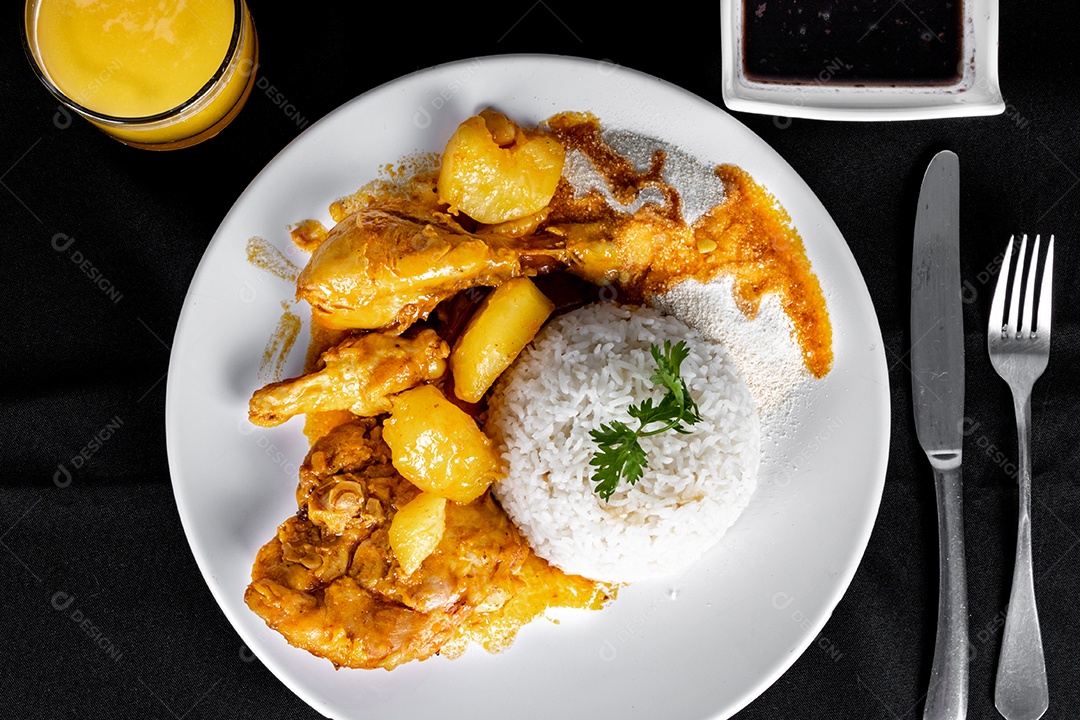 Ensopado de frango caseiro com tomate, cebola e batata em uma mesa de tecido preto e prato com arroz, feijão e suco de laranja. Vista do topo.