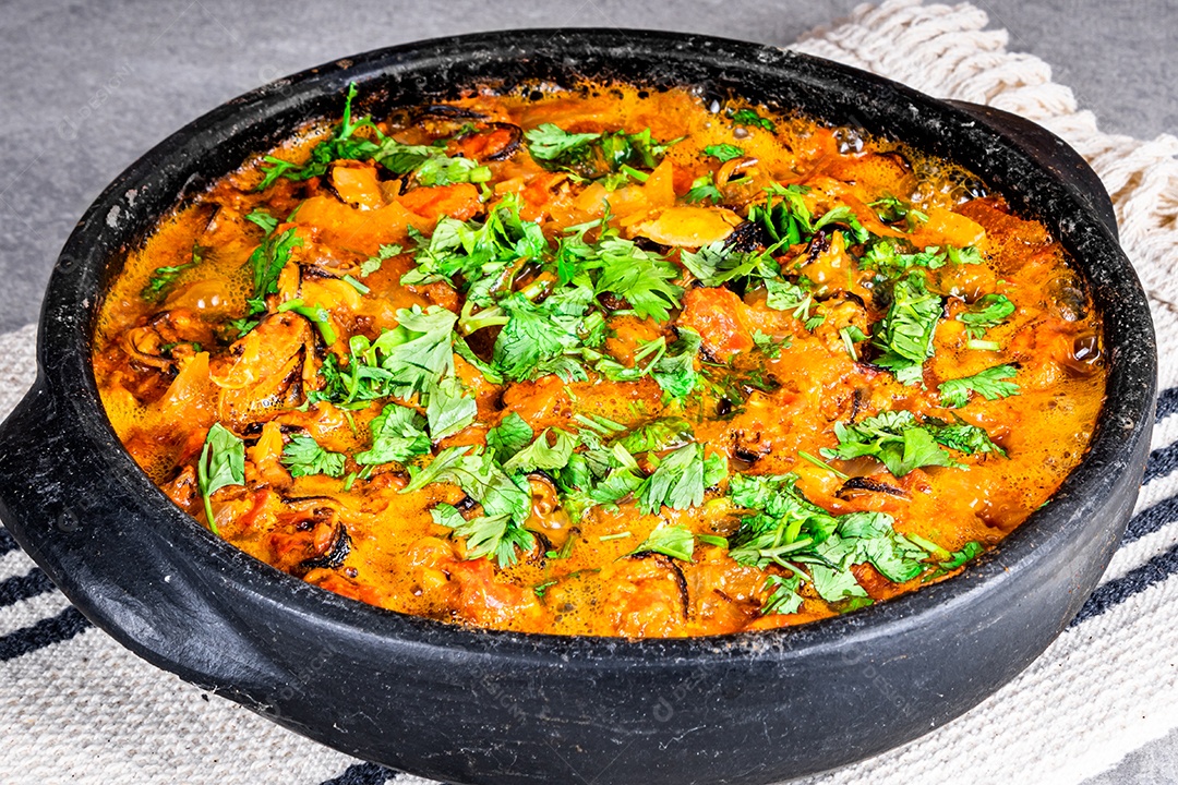 Deliciosa moqueca de peixe brasileiro com tomate, cebola, azeite, coentro e semente de orocum. Feito em panela de barro sobre mesa branca e fundo preto