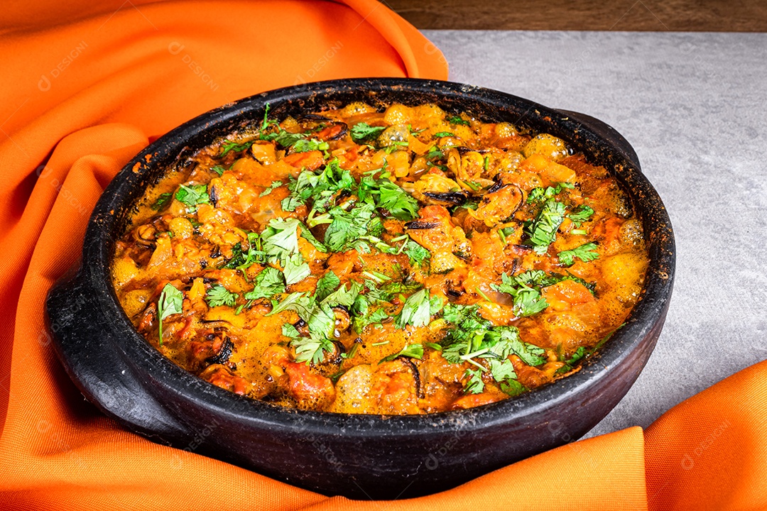 Deliciosa moqueca de peixe brasileiro com tomate, cebola, azeite, coentro e semente de orocum. Feito em panela de barro sobre mesa branca e fundo preto