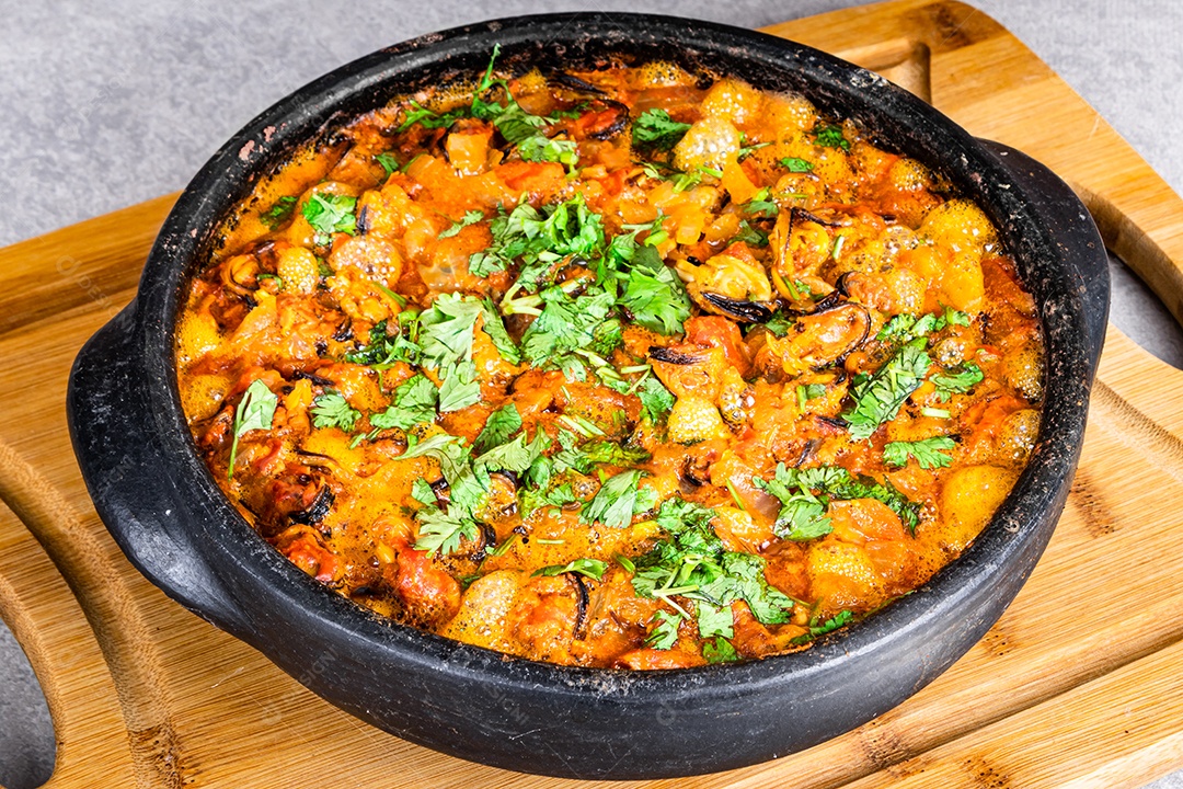 Deliciosa moqueca de peixe brasileiro com tomate, cebola, azeite, coentro e semente de orocum. Feito em panela de barro sobre mesa branca e fundo preto