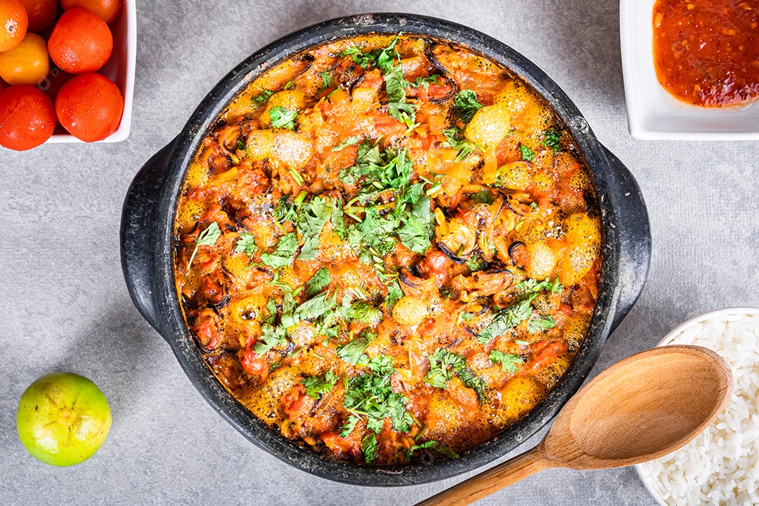 Deliciosa moqueca de peixe brasileiro com tomate, cebola, azeite, coentro e semente de orocum. Feito em panela de barro sobre mesa branca e fundo preto. Com arroz.