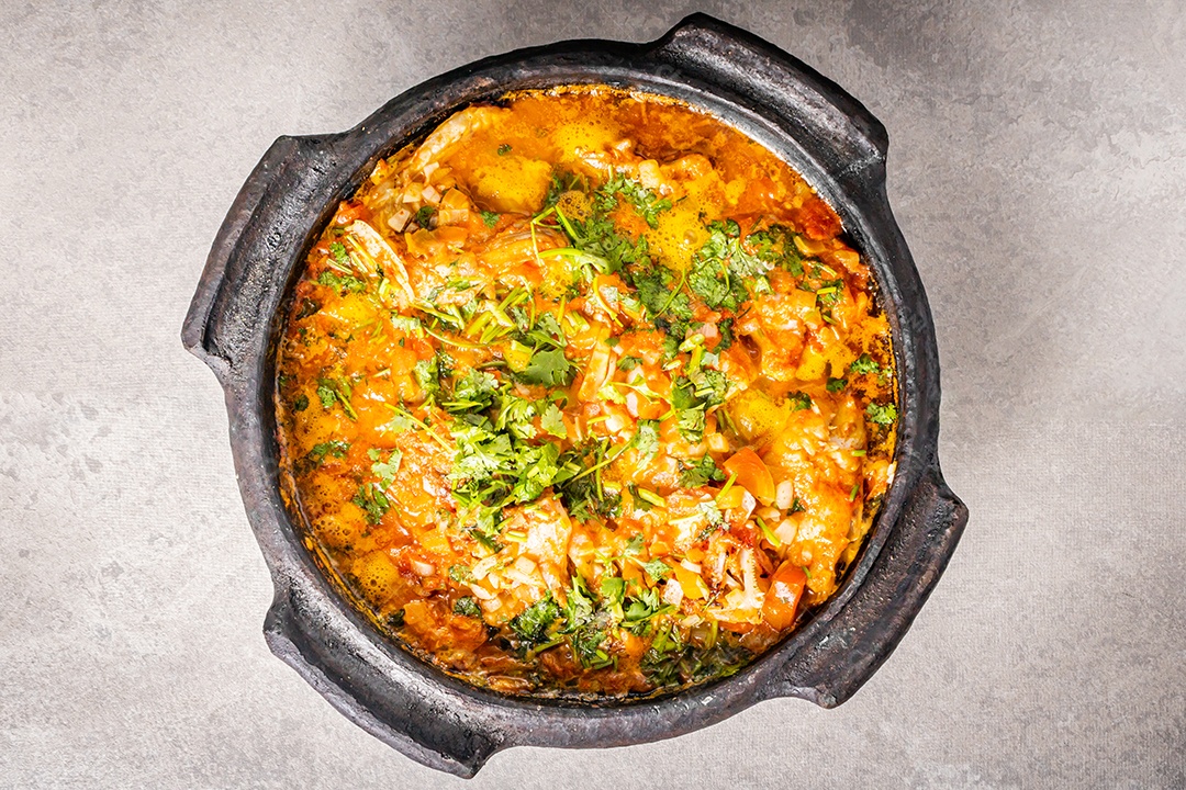 Deliciosa moqueca de peixe brasileiro com tomate, cebola, azeite, coentro e semente de orocum. Feito em panela de barro sobre mesa branca e fundo preto. Com arroz.