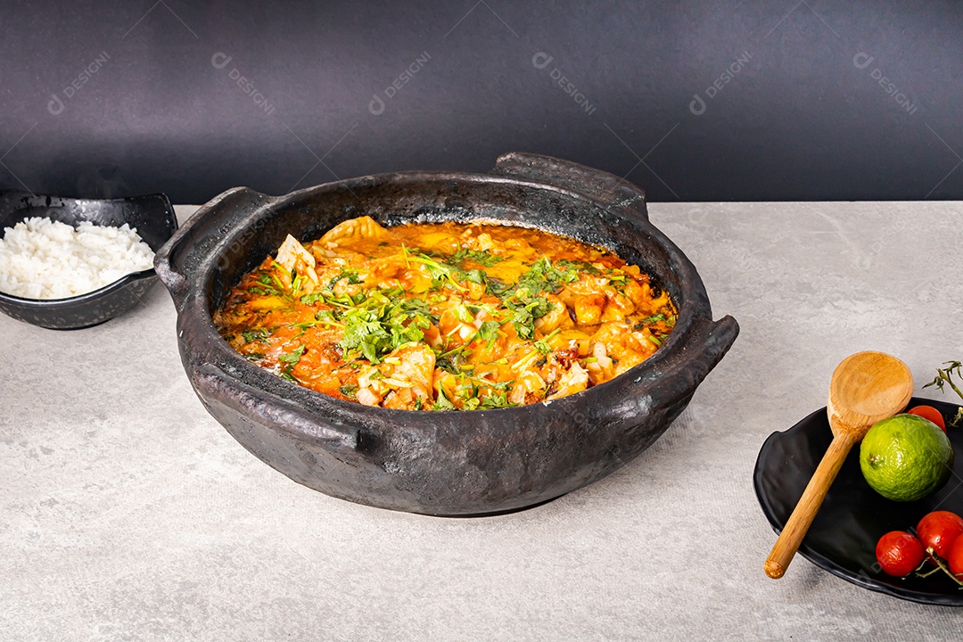 Deliciosa moqueca de peixe brasileiro com tomate, cebola, azeite, coentro e semente de orocum. Feito em panela de barro sobre mesa branca e fundo preto. Com arroz.