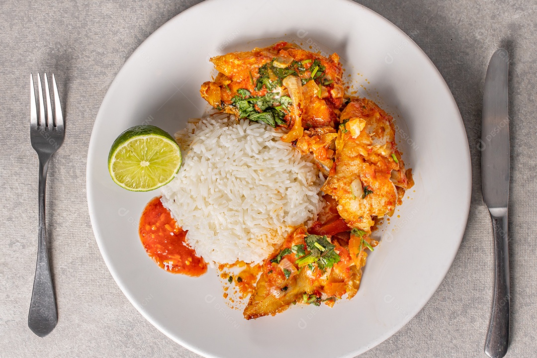 Deliciosa moqueca de peixe brasileiro com tomate, cebola, azeite, coentro e semente de orocum. Servido em prato de porcelana branca com arroz. Com toalha laranja ao redor.