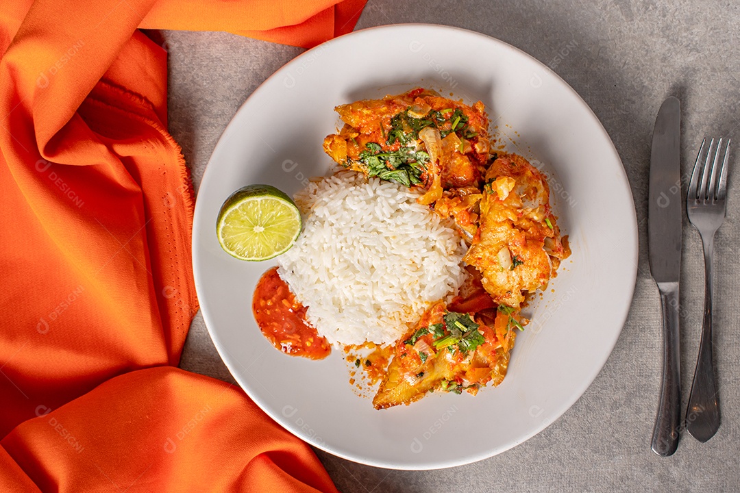 Deliciosa moqueca de peixe brasileiro com tomate, cebola, azeite, coentro e semente de orocum. Servido em prato de porcelana branca com arroz. Com toalha laranja ao redor.