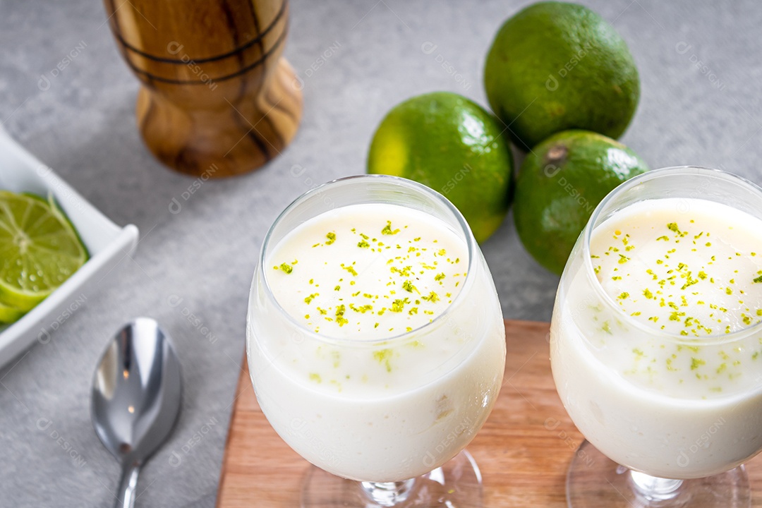 Deliciosa mousse de limão sobre uma tábua de madeira e um fundo de mesa branco, com limões inteiros e fatiados ao fundo e uma colher de metal