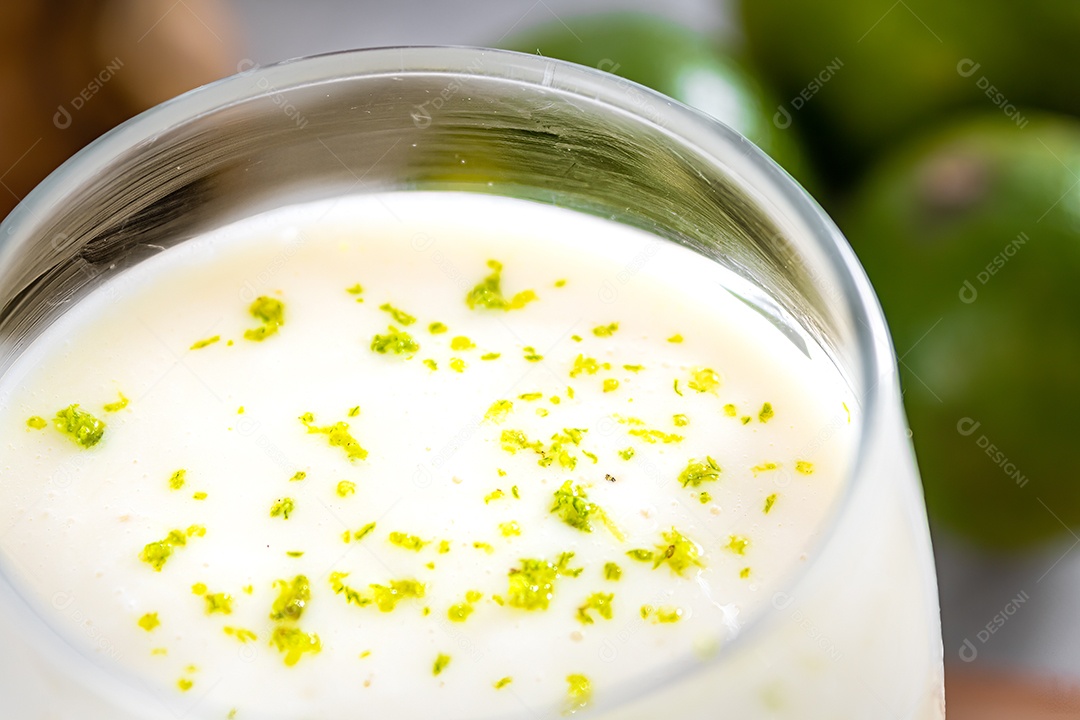 Deliciosa mousse de limão sobre uma tábua de madeira e um fundo de mesa branco, com limões inteiros e fatiados ao fundo e uma colher de metal