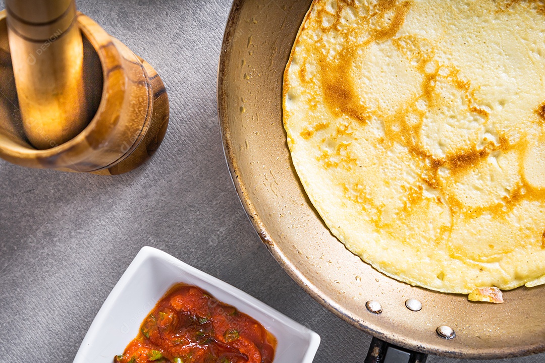 Deliciosas panquecas em uma frigideira sobre uma mesa branca, com trigo, ovos, azeite, molho de tomate e uma toalha xadrez ao fundo