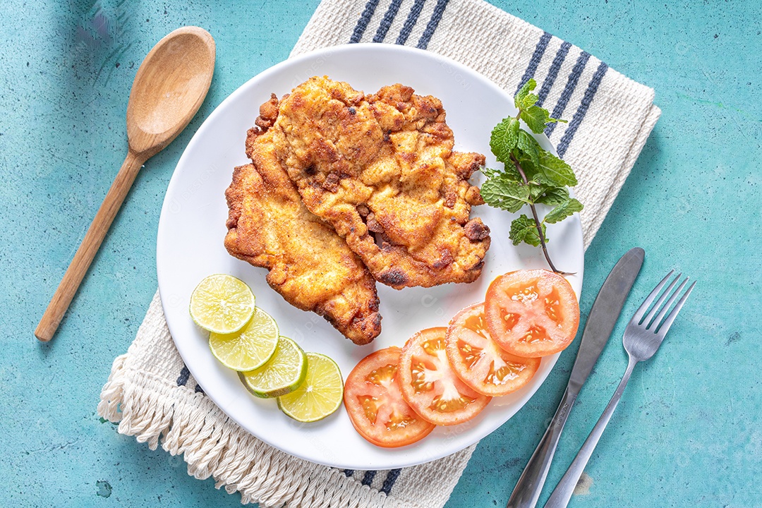 Delicioso frango à milanesa (frango empanado com farinha de rosca e ovo), prato típico brasileiro. Em um prato branco com tomate, limão no mármore azul. Detalhe do bife cortado ao meio.