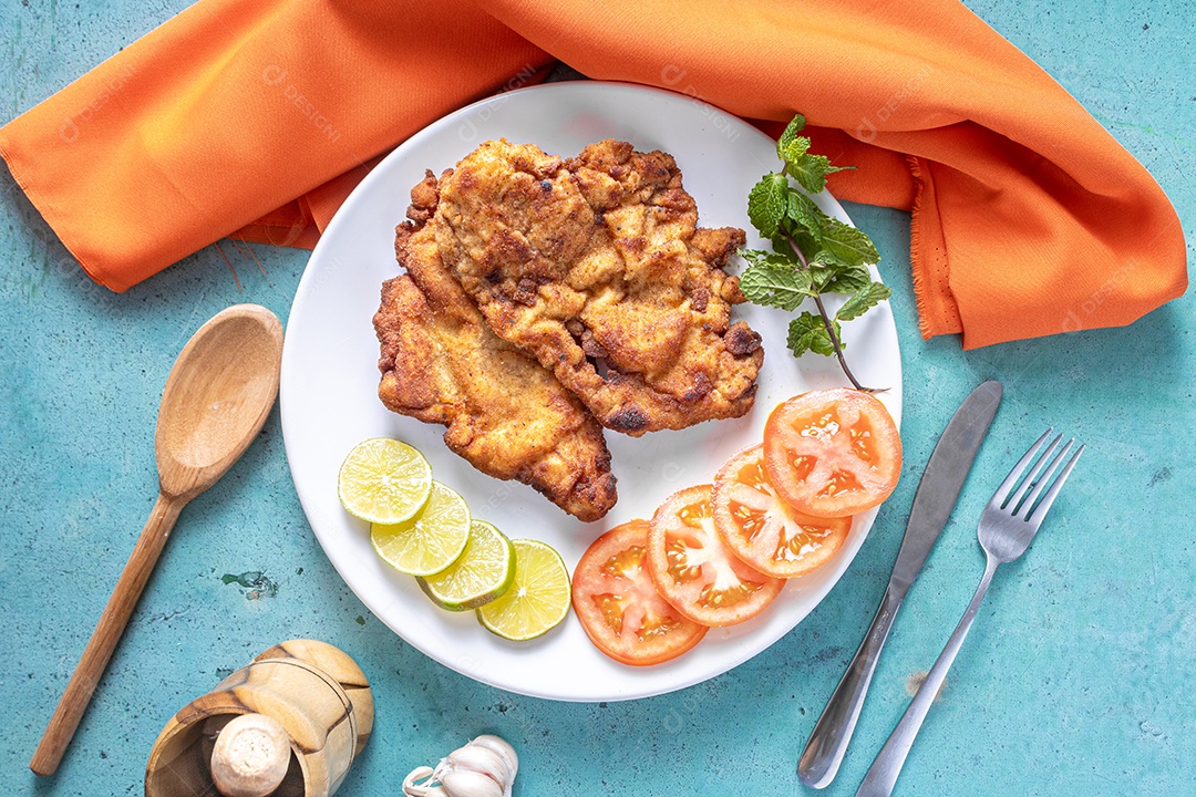 Delicioso frango à milanesa (frango empanado com farinha de rosca e ovo), prato típico brasileiro. Em um prato branco com tomate, limão no mármore azul. Detalhe do bife cortado ao meio.