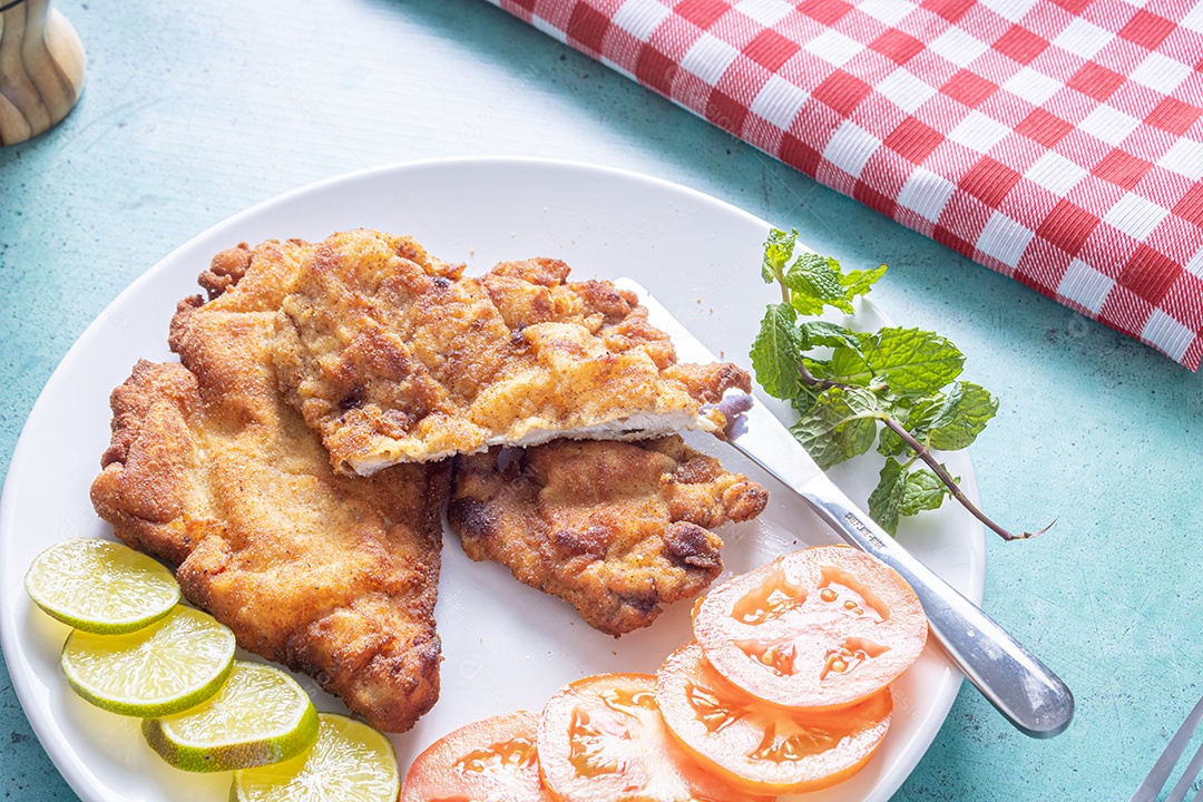 Delicioso frango à milanesa (frango empanado com farinha de rosca e ovo), prato típico brasileiro. Em um prato branco com tomate, limão no mármore azul. Detalhe do bife cortado ao meio.