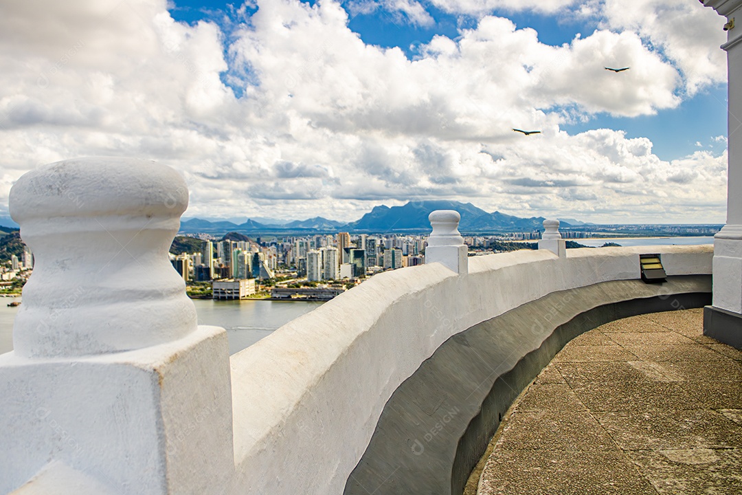 Detalhe de parapeito branco com linha curva, mostrando a cidade de Vitória e pássaros no céu ao fundo.