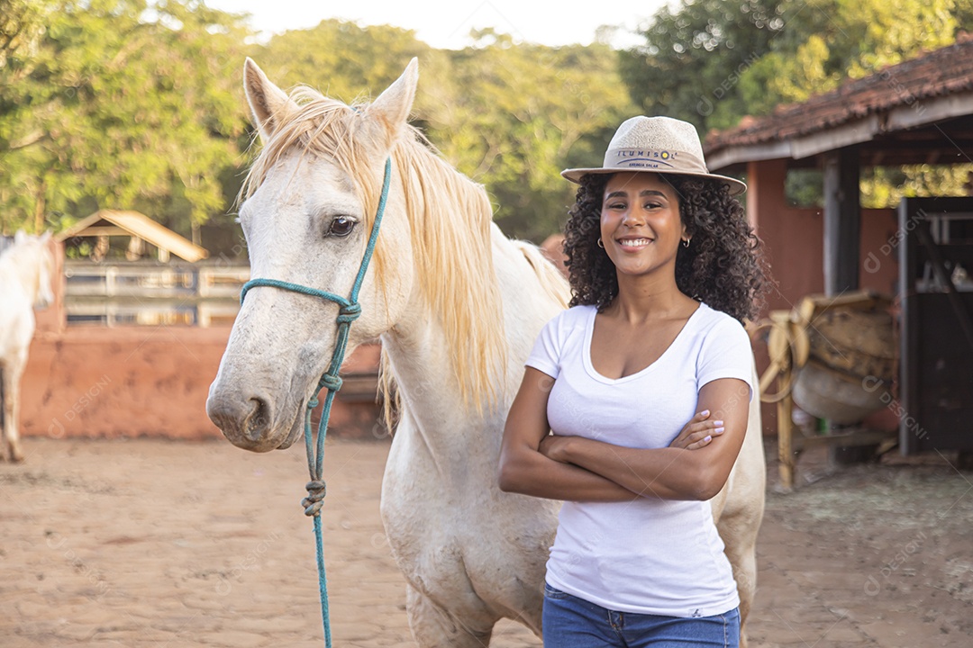 Mulher jovem feliz e sorridente sobre fazenda ao lado de seu cavalo