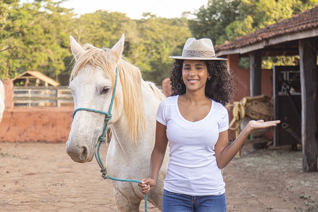 Mulher jovem feliz e sorridente sobre fazenda ao lado de seu cavalo