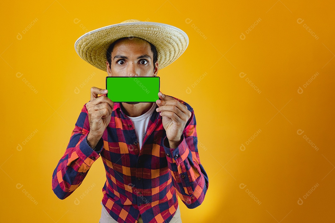 Homem negro com roupa de festa junina segurando um celular na frente de uma boca isolada em fundo amarelo.