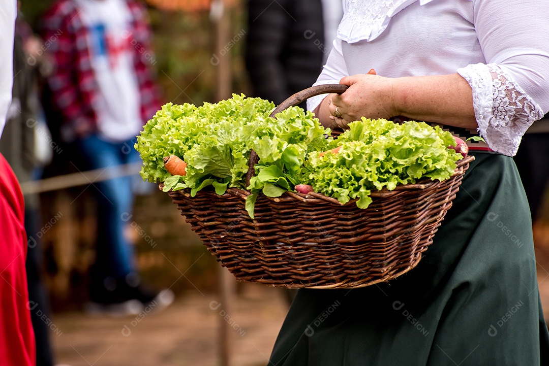 Mulher desfilando com roupas velhas segurando cesto com legumes, simbolizando a chegada de imigrantes