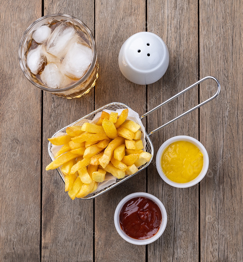 Batatas fritas em uma cesta com sal, ketchup, mostarda e um copo de refrigerante sobre a mesa de madeira