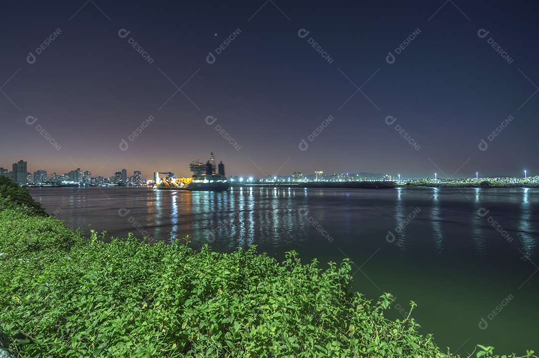 Navio cargueiro navegando no mar à noite com a cidade de Itajaí-SC ao fundo e luzes acesas, foto em longa exposição.
