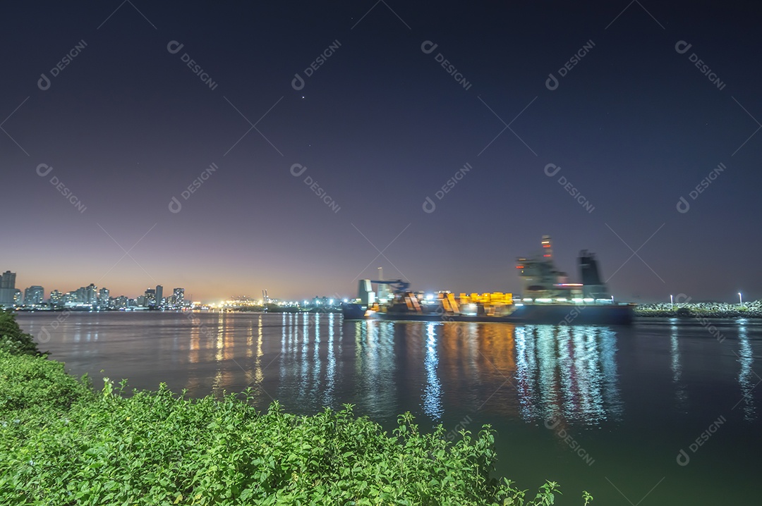 Navio cargueiro navegando no mar à noite com a cidade de Itajaí-SC ao fundo e luzes acesas, foto em longa exposição.