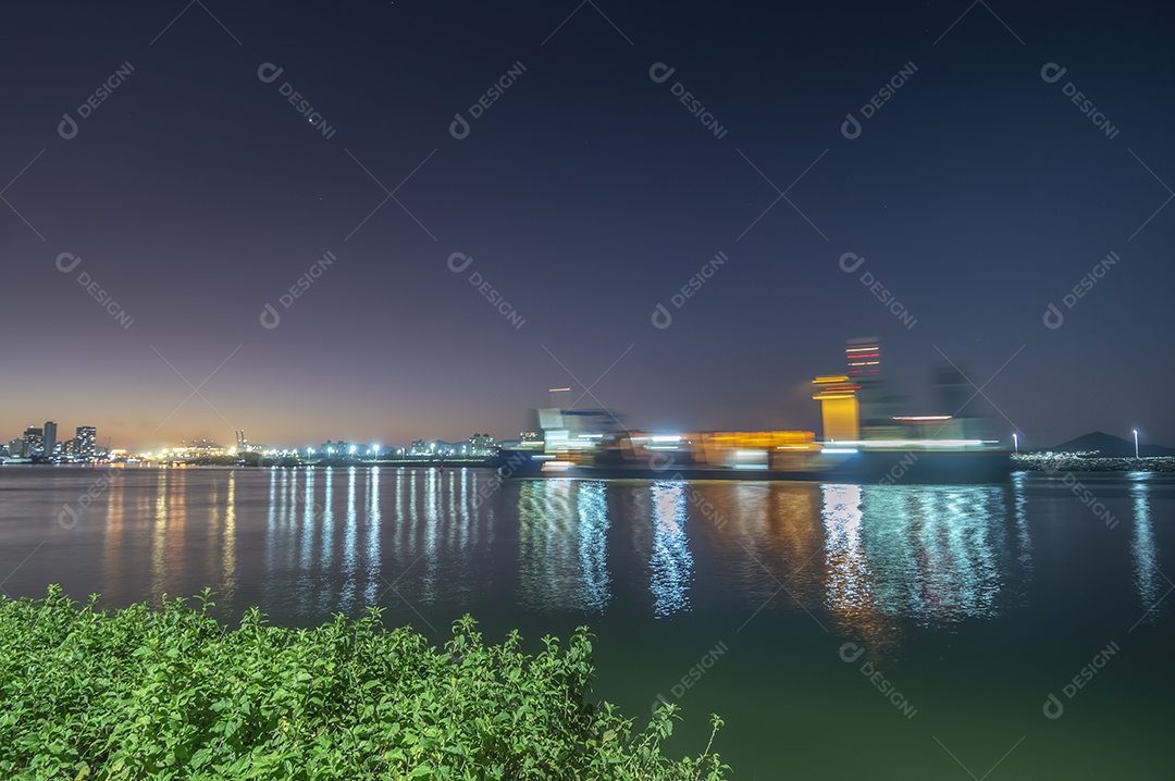 Navio cargueiro navegando no mar à noite com a cidade de Itajaí-SC ao fundo e luzes acesas, foto em longa exposição.