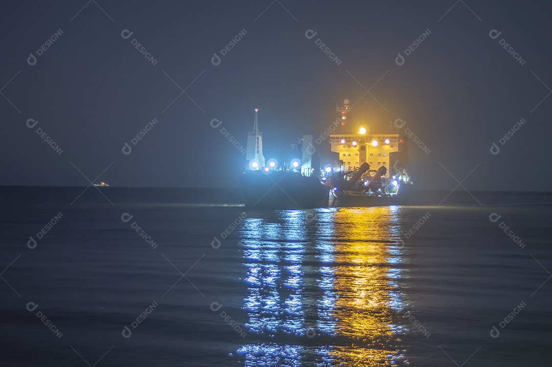 Navio cargueiro navegando no mar à noite com a cidade de Itajaí-SC ao fundo e luzes acesas, foto em longa exposição.
