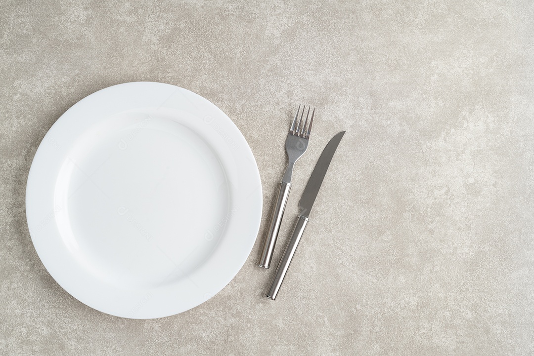 Placa cerâmica vazia branca com garfo e faca sobre fundo de pedra com espaço de cópia.
