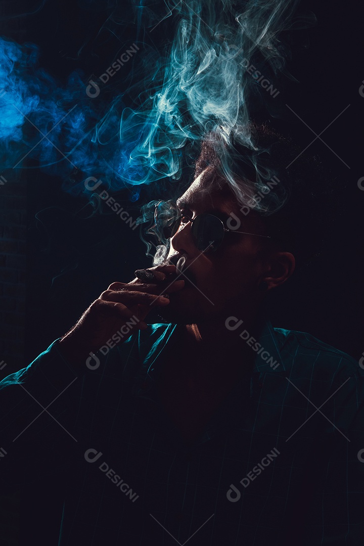 Jovem negro com óculos fumando charuto com neblina no fundo preto, luz baixa.