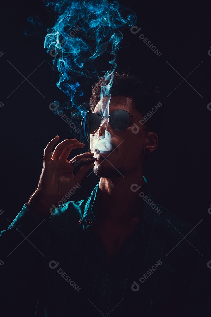 Jovem negro com óculos fumando charuto com neblina no fundo preto, luz baixa.