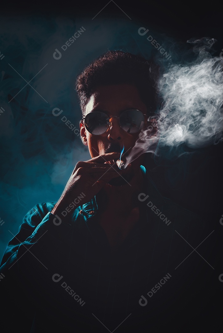 Jovem negro com óculos fumando charuto com neblina no fundo preto, luz baixa.
