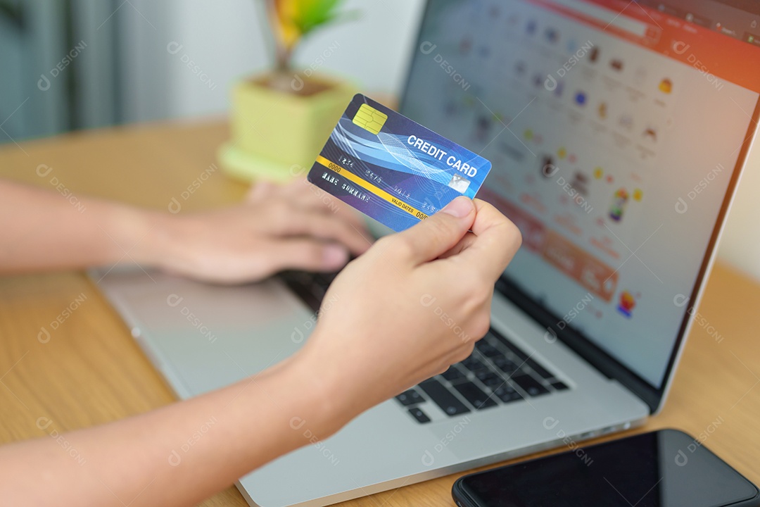 mão de uma mulher segurando o cartão de crédito e usando o laptop com o celular para fazer compras on-line enquanto faz o pedido.