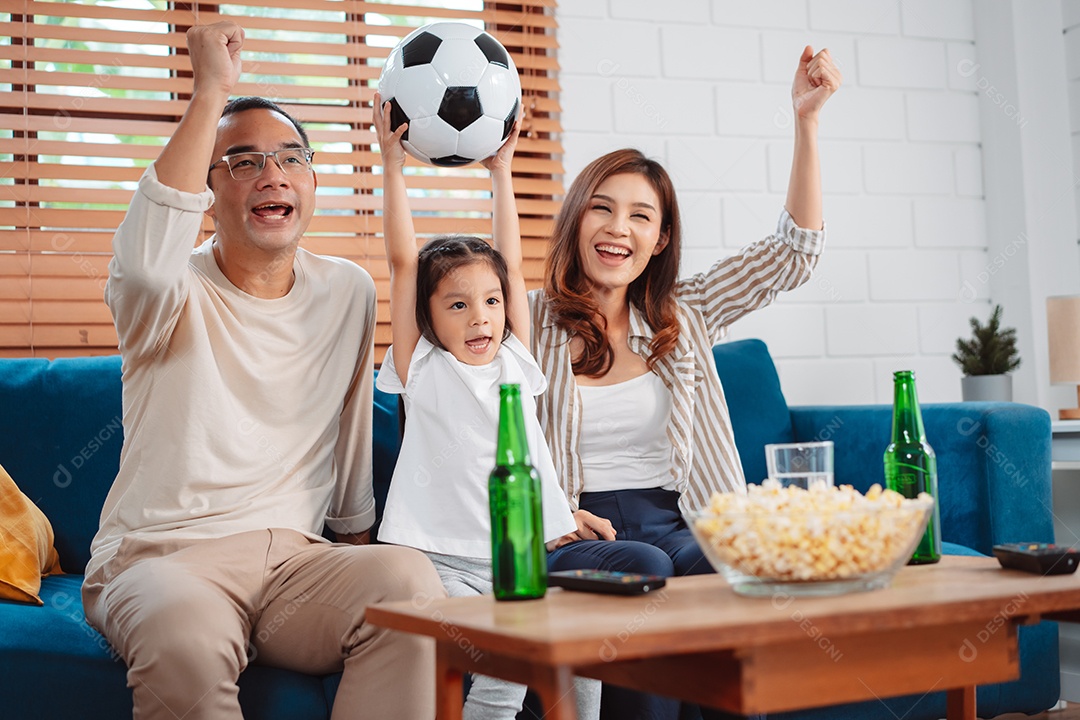 Família asiática com a filha assistindo futebol esportivo torcendo animado comendo pipoca no sofá na sala