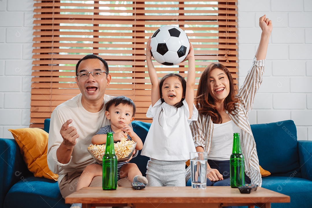 Família asiática com a filha assistindo futebol esportivo torcendo animado comendo pipoca no sofá na sala