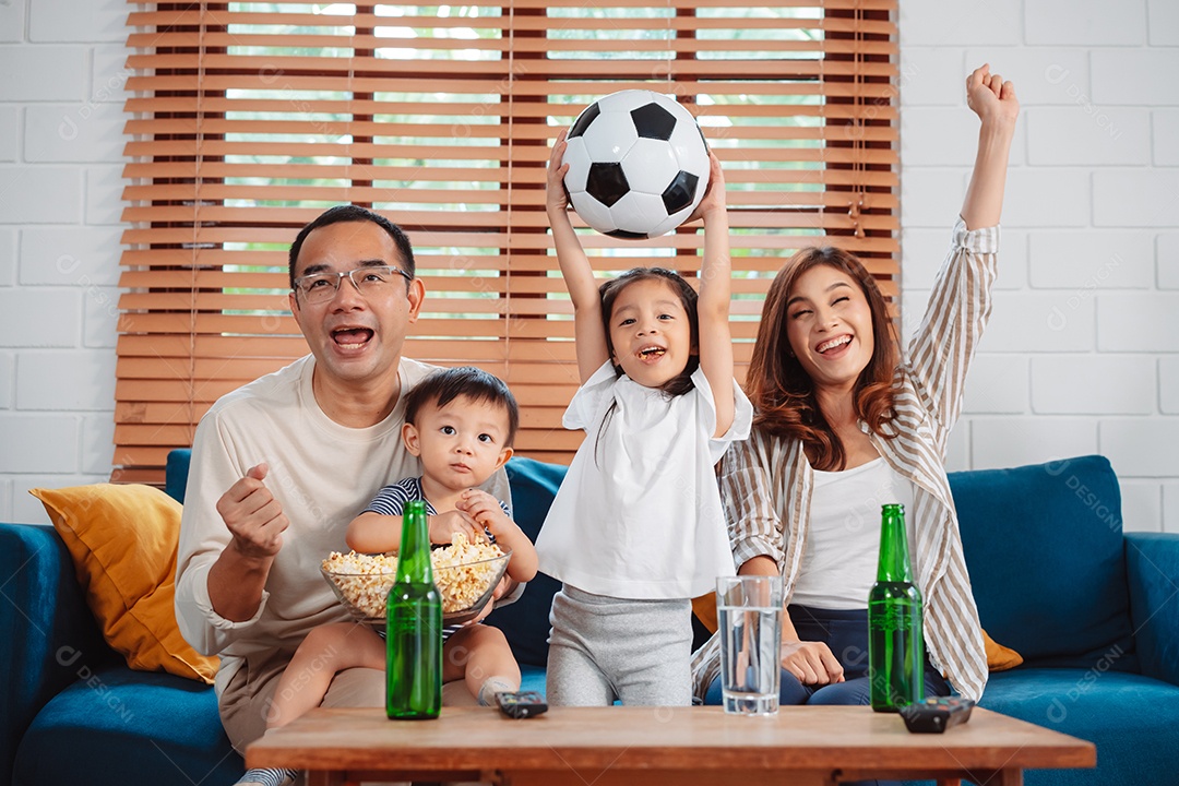 Família asiática com a filha assistindo futebol esportivo torcendo animado comendo pipoca no sofá na sala