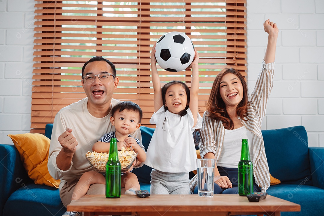Família asiática com a filha assistindo futebol esportivo torcendo animado comendo pipoca no sofá na sala
