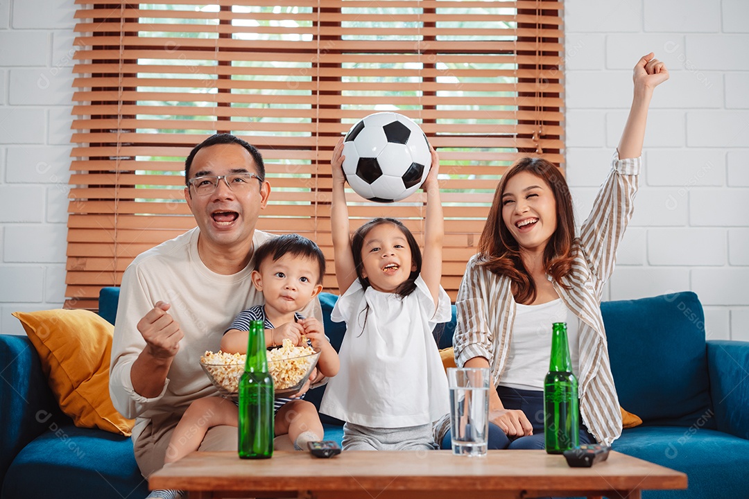 Família asiática com a filha assistindo futebol esportivo torcendo animado comendo pipoca no sofá na sala