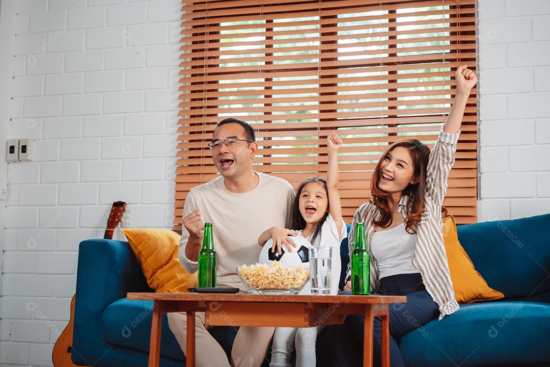 Família asiática com a filha assistindo futebol esportivo torcendo animado comendo pipoca no sofá na sala