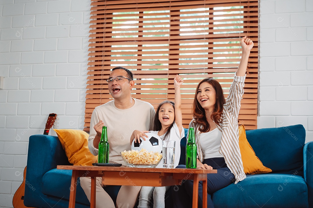 Família asiática com a filha assistindo futebol esportivo torcendo animado comendo pipoca no sofá na sala