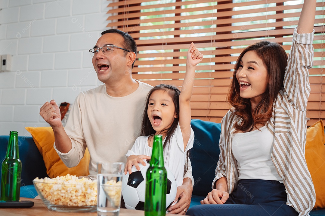 Família asiática com a filha assistindo futebol esportivo torcendo animado comendo pipoca no sofá na sala