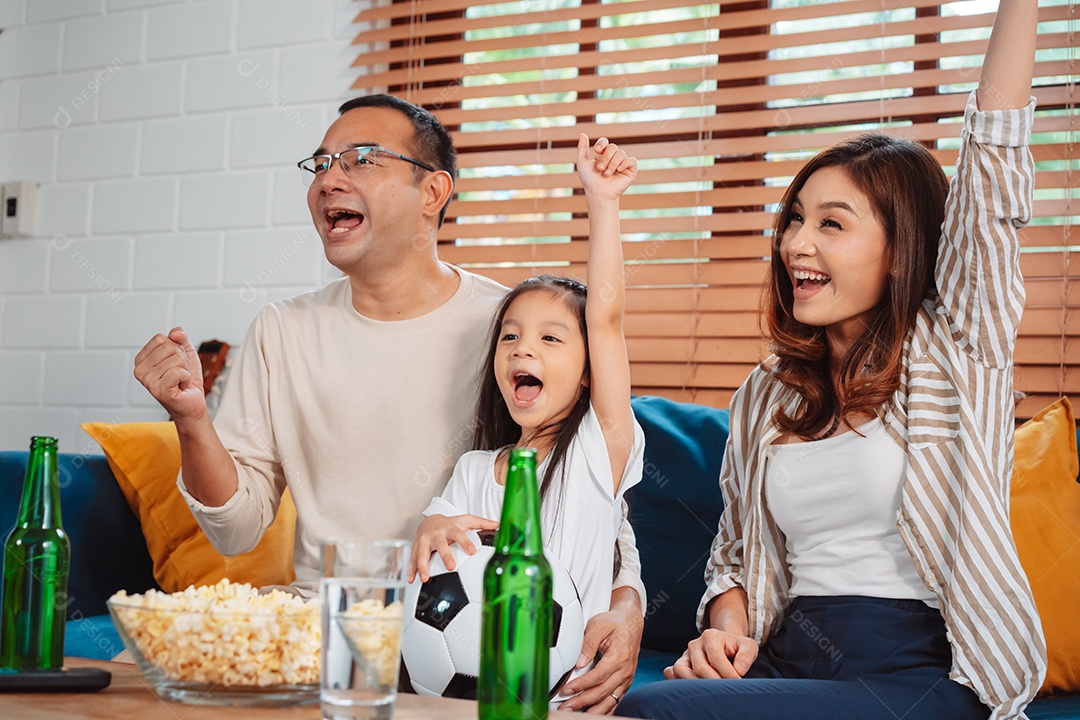 Família asiática com a filha assistindo futebol esportivo torcendo animado comendo pipoca no sofá na sala