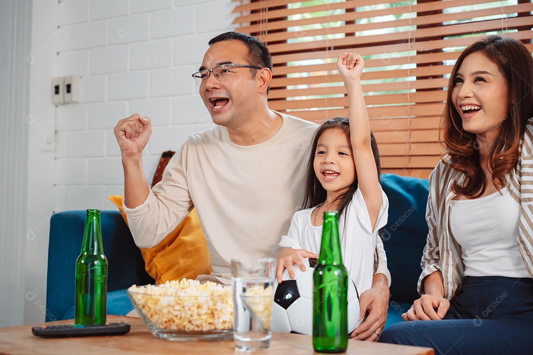 Família asiática com a filha assistindo futebol esportivo torcendo animado comendo pipoca no sofá na sala