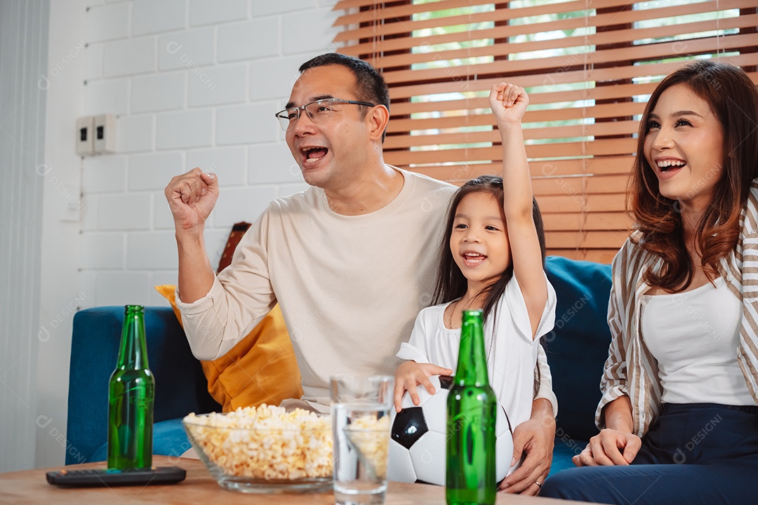 Família asiática com a filha assistindo futebol esportivo torcendo animado comendo pipoca no sofá na sala