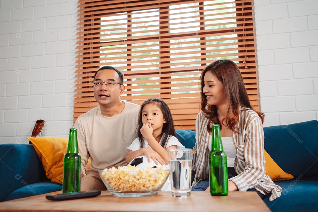 Família asiática com a filha assistindo futebol esportivo torcendo animado comendo pipoca no sofá na sala