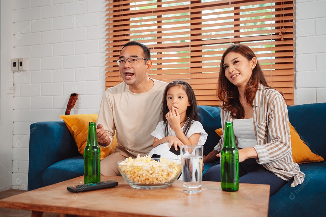 Família asiática com a filha assistindo futebol esportivo torcendo animado comendo pipoca no sofá na sala
