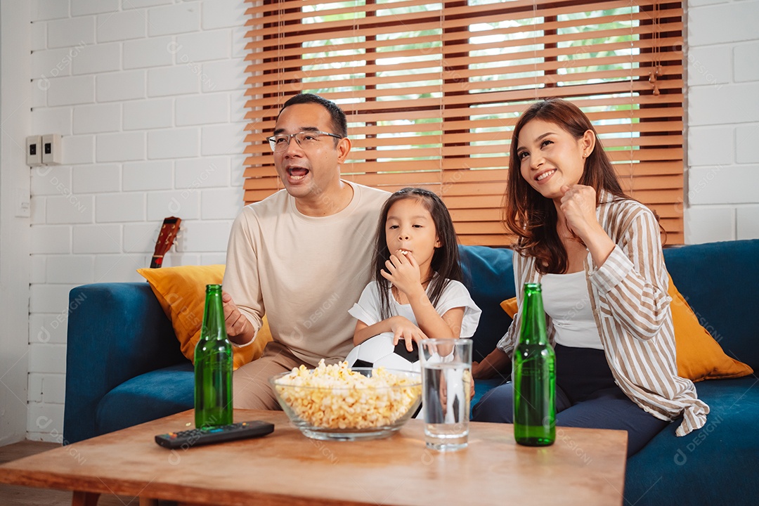 Família asiática com a filha assistindo futebol esportivo torcendo animado comendo pipoca no sofá na sala