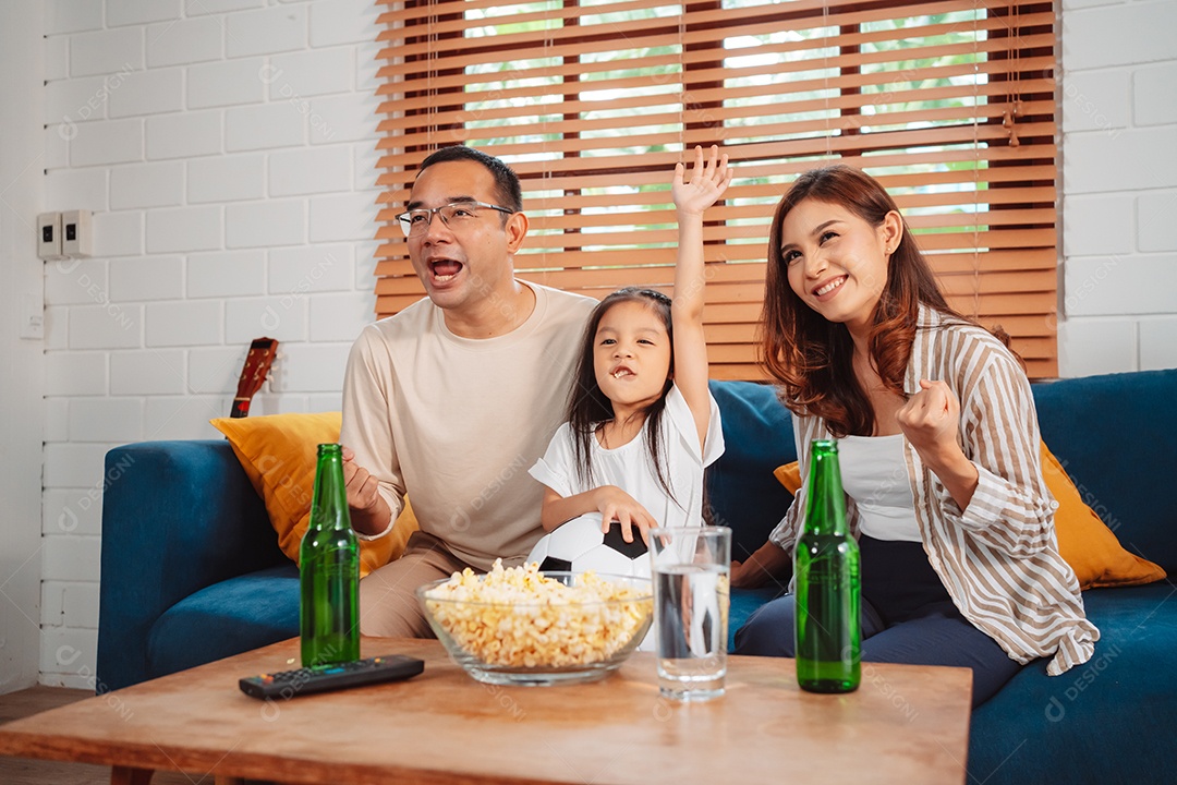 Família asiática com a filha assistindo futebol esportivo torcendo animado comendo pipoca no sofá na sala